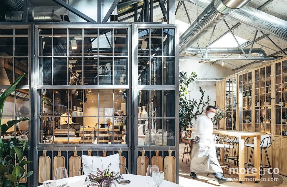 Un moderno interior de restaurante de estilo industrial con grandes ventanales, muebles de madera, conductos a la vista y una persona vestida de blanco caminando por el espacio.