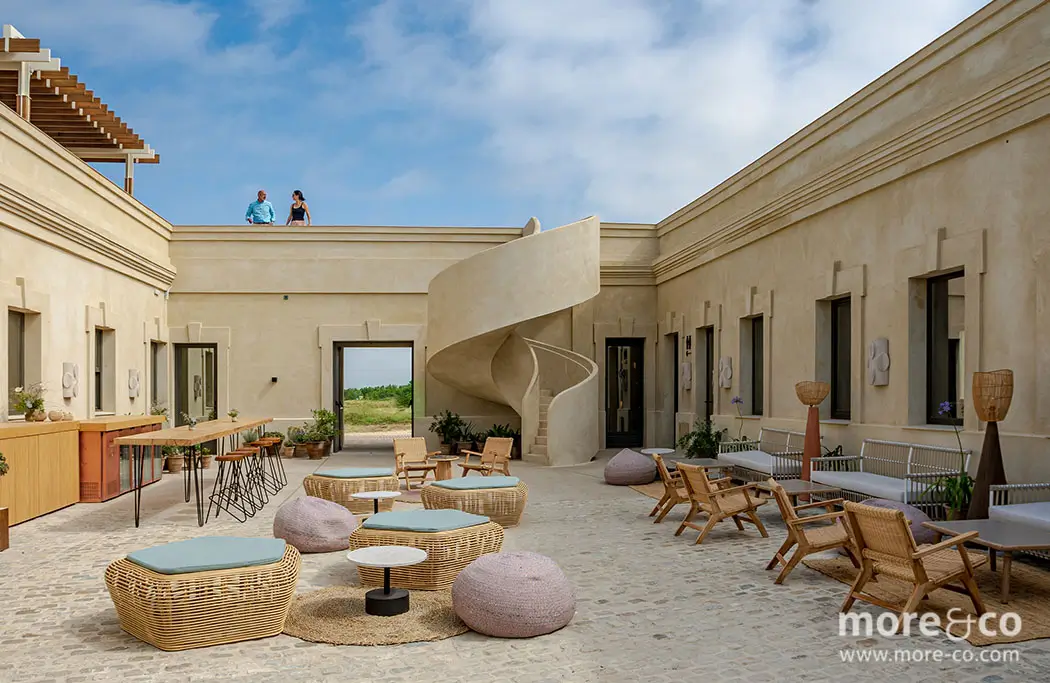 Patio abierto con muebles de salón de mimbre, mesas circulares y pufs sobre un suelo empedrado; una escalera de caracol conduce a una azotea donde hay dos personas de pie.
