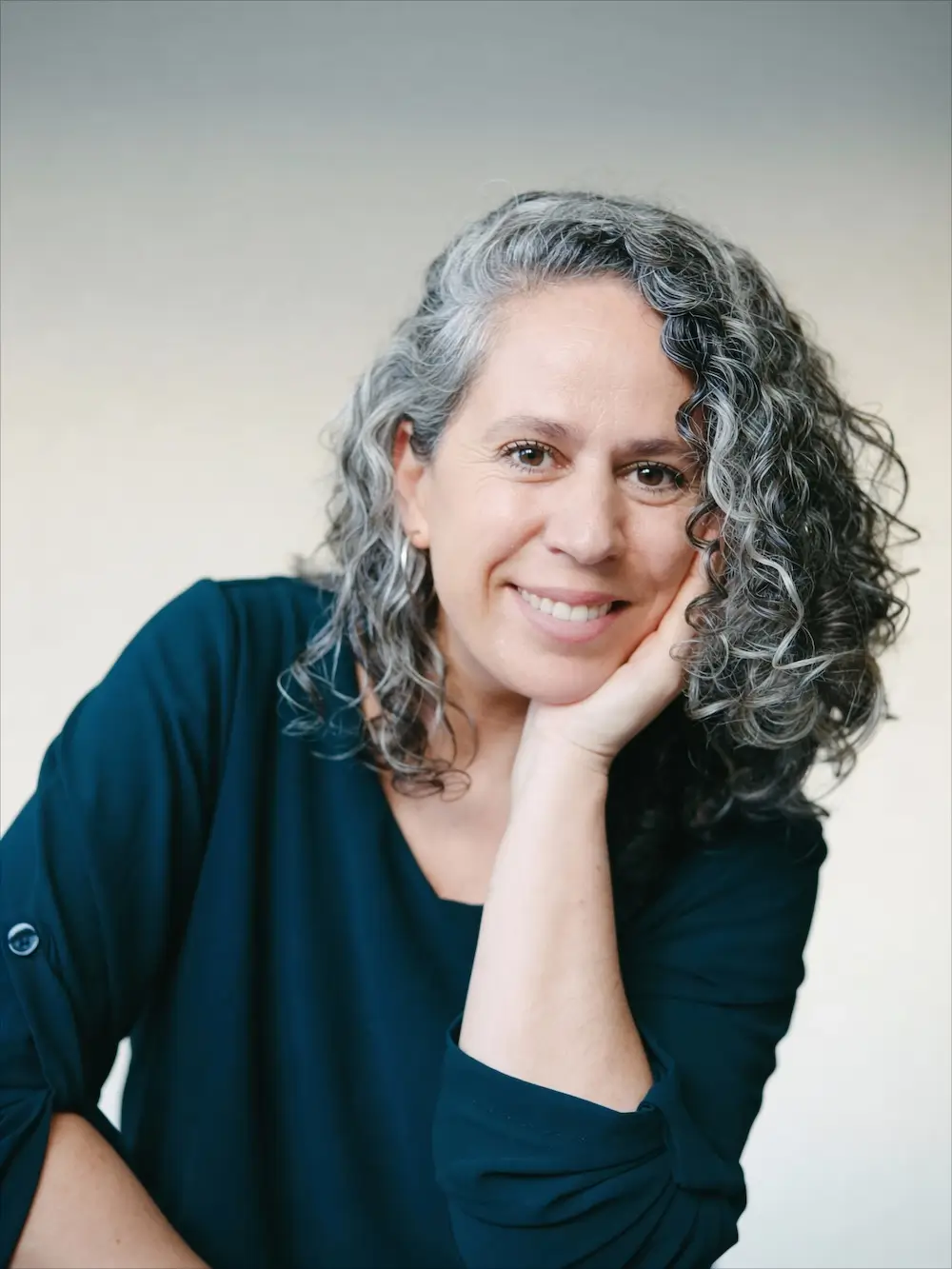 Mujer con el pelo rizado y gris, vestida con un top azul oscuro, sonriendo y apoyando la cabeza en la mano, sobre un fondo claro.