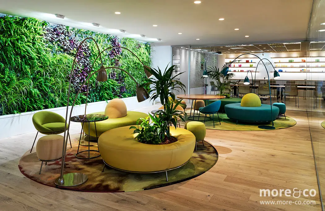 Moderno salón de oficina con jardín vertical verde, coloridos asientos circulares, mesas redondas y lámparas de pie sobre suelo de madera. Una pared de cristal separa el salón de la zona de reuniones.