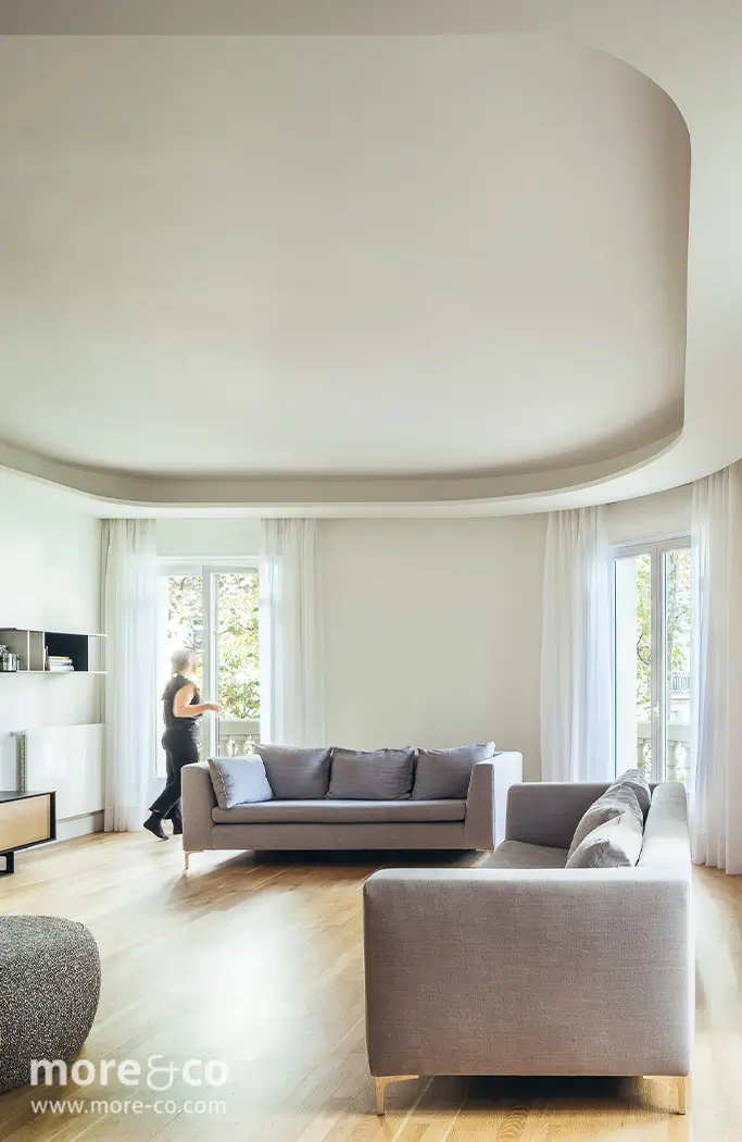 Un salón moderno con dos sofás grises, suelo de madera clara, grandes ventanales con cortinas transparentes y una persona de pie cerca de la ventana.